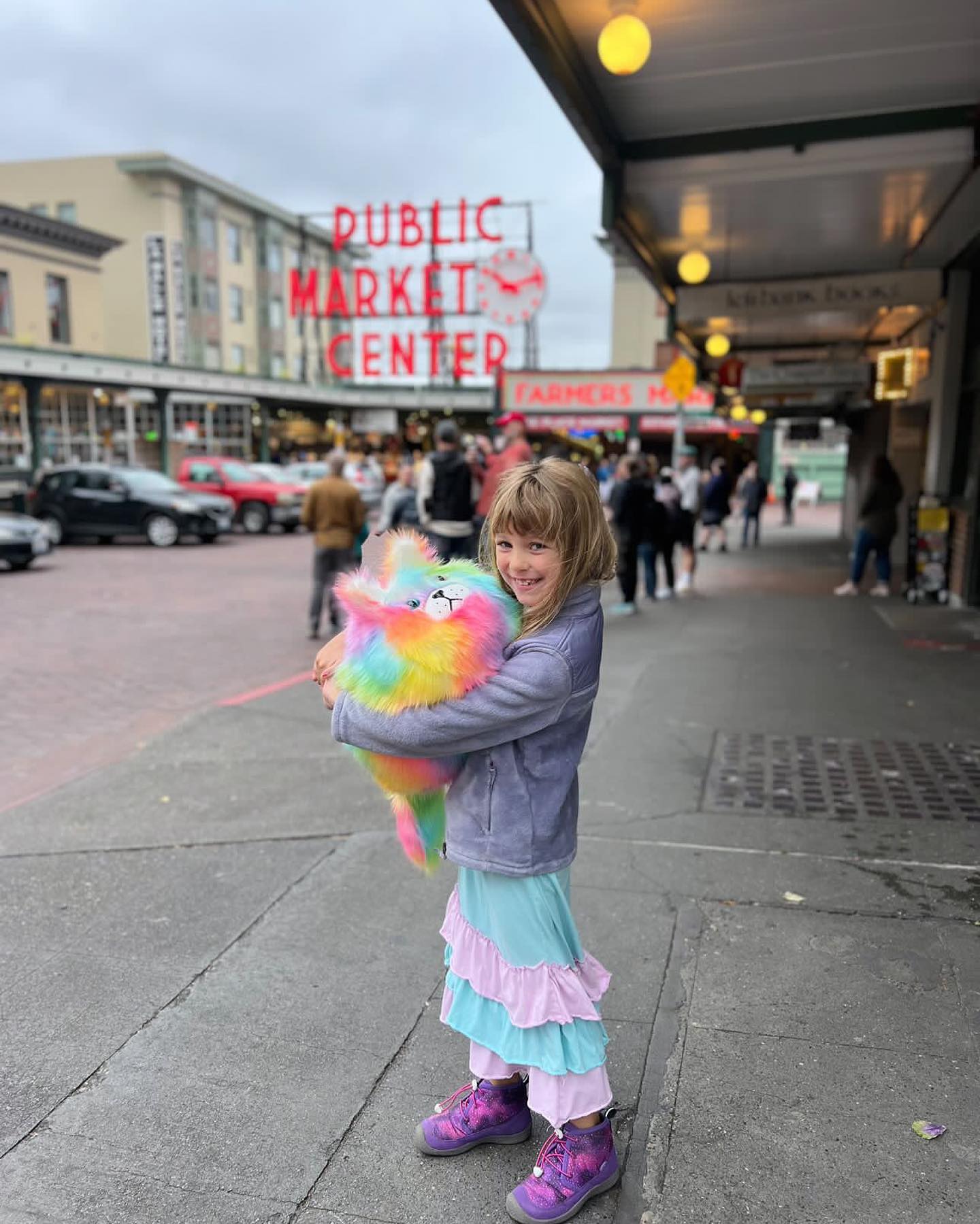 A girl hugs a large rainbow colored cat loaf stuffed animal in front of the historic pike place market neon sign.