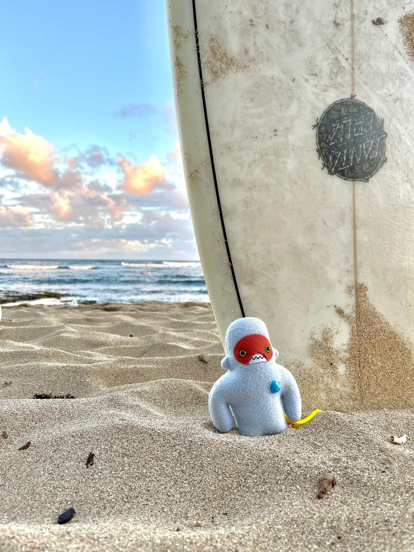 A small light blue yeti plush animal stands in the sand at the beach with a huge surfboard backdrop.