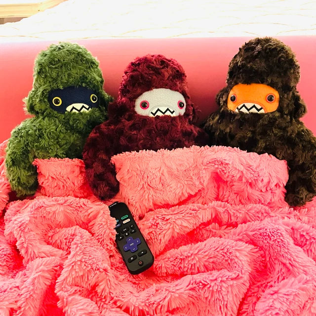 Threen plush sasquatch are nestled under a pink blanket with remote within reach of one plush paw.