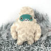 Amidst the frosted pine branches stands a handmade plush monster, this furry creature boasts a cream colored, curly texture. Its teal face, big orange eyes, and toothy grin make it the perfect yeti sidekick for any cozy quest adventure.