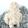 Back view of a plush monster with curly cream colored fur stands in front of some white foliage.