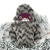 Amidst the frosted pine branches stands a handmade plush monster, this furry creature boasts a gray, curly texture. Its dark purple face, big green eyes, and toothy grin make it the perfect yeti sidekick for any cozy quest adventure.