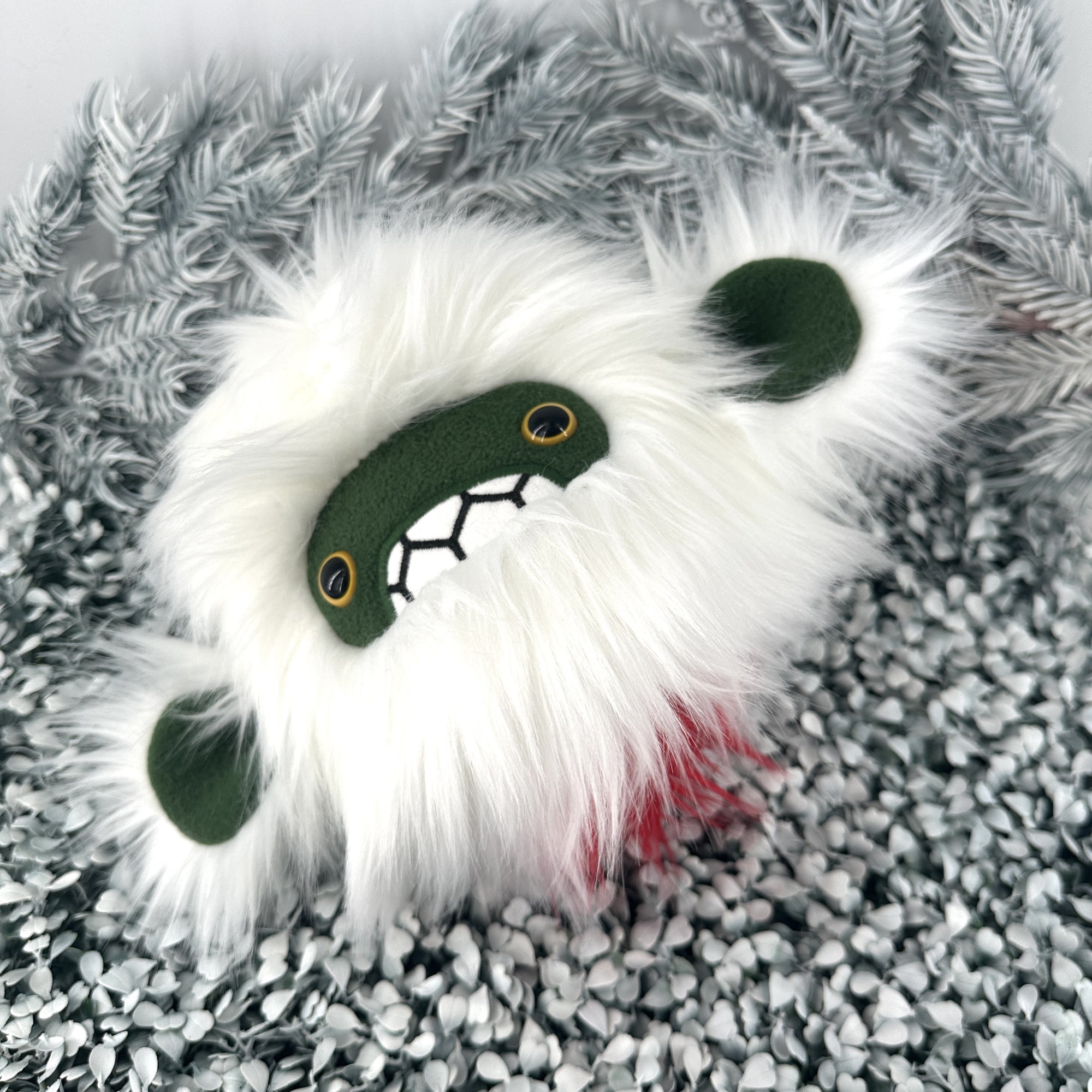 A plush toy resembling a white yeti with a fluffy faux fur head, dark face, and expressive eyes lies on a textured, pebble-like surface. A tiny bit of red fur is visible on the underside.