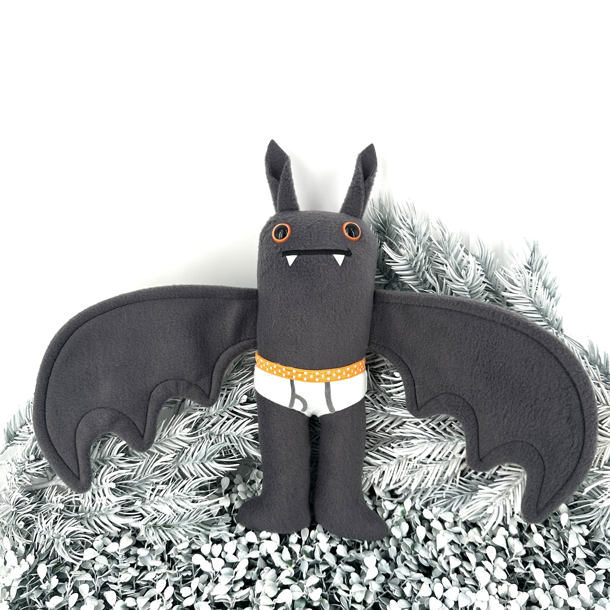 A plush Flasher Bat with orange eyes, small fangs, and tiny tighty whities stands on bed of frosty pine branches. 