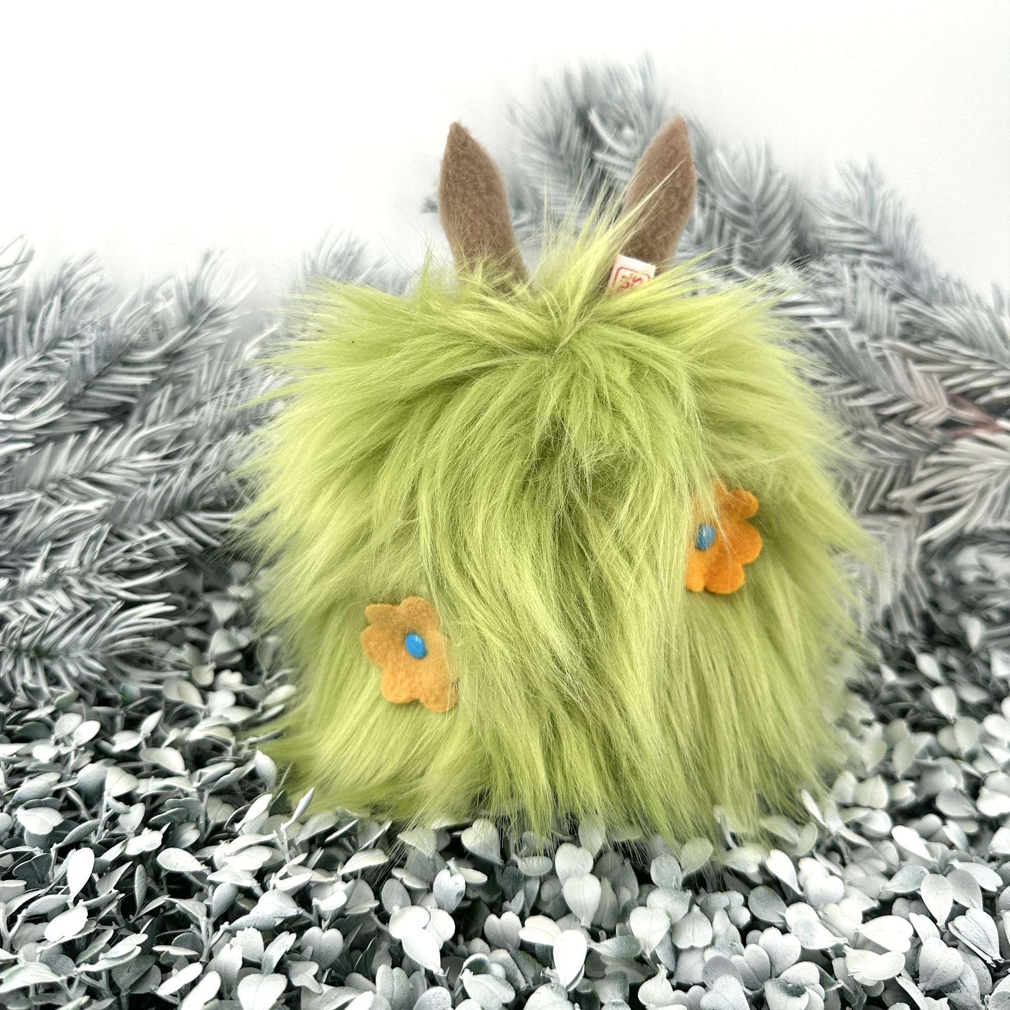 Bright green faux fur creature with yellow fleece flowers in its hair sits on white foliage.