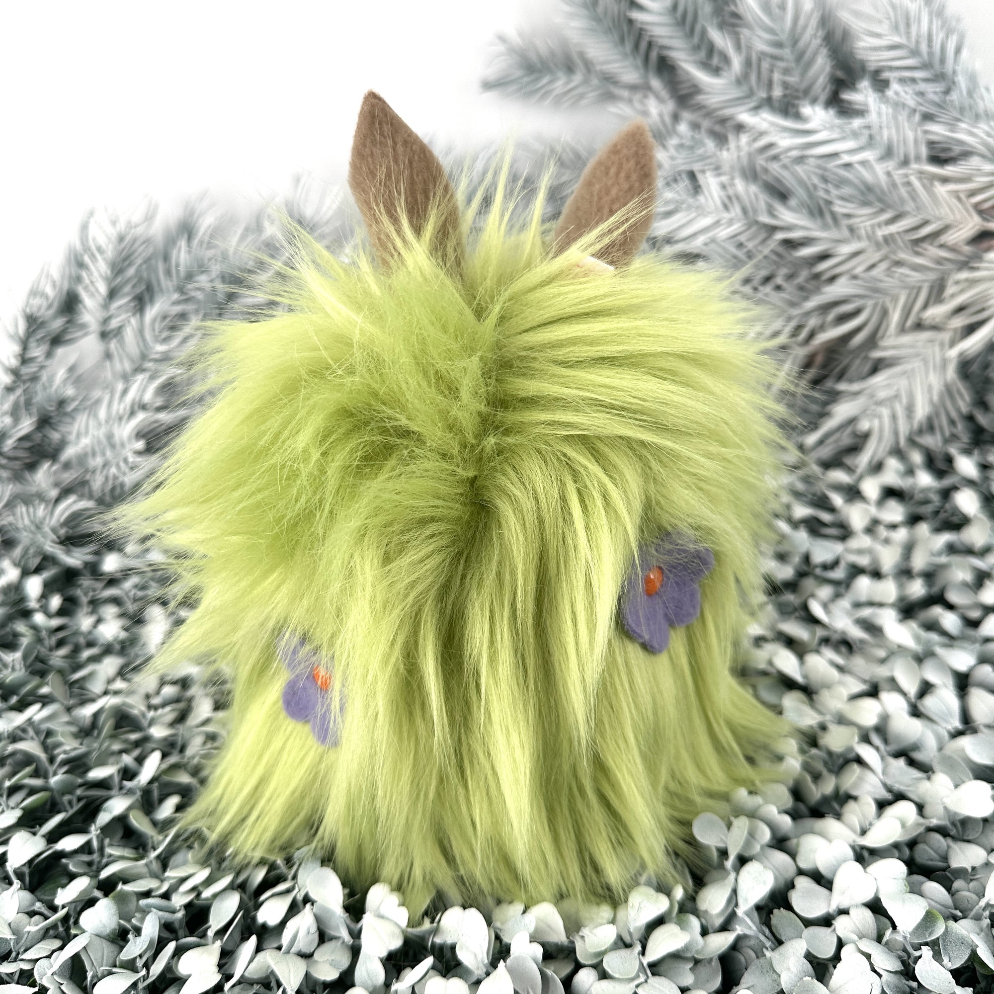 Bright green faux fur creature with purple fleece flowers in its hair sits on white foliage.