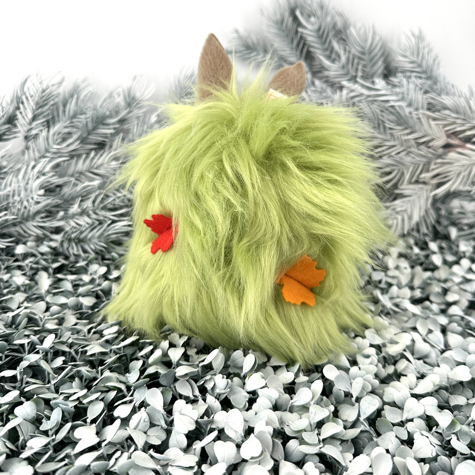 Bright green faux fur creature with colorful felt leaves in its hair sits on white foliage.