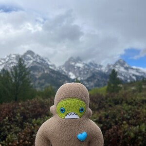 Small tan fleece sasquatch plush stands in front of mountain backdrop.