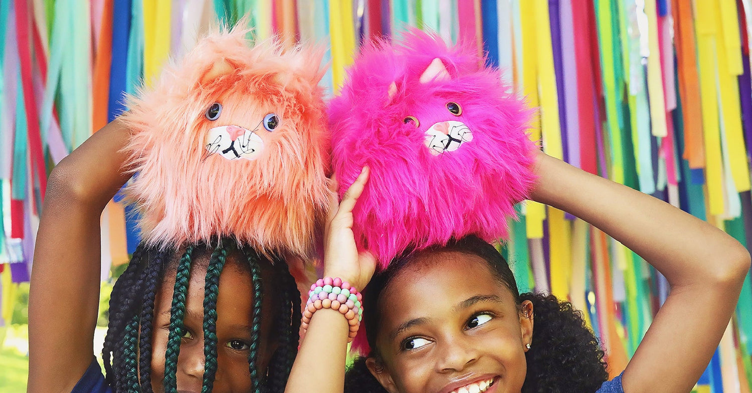 Two girls shown holding furry cats on top of their heads with colorful striped background.