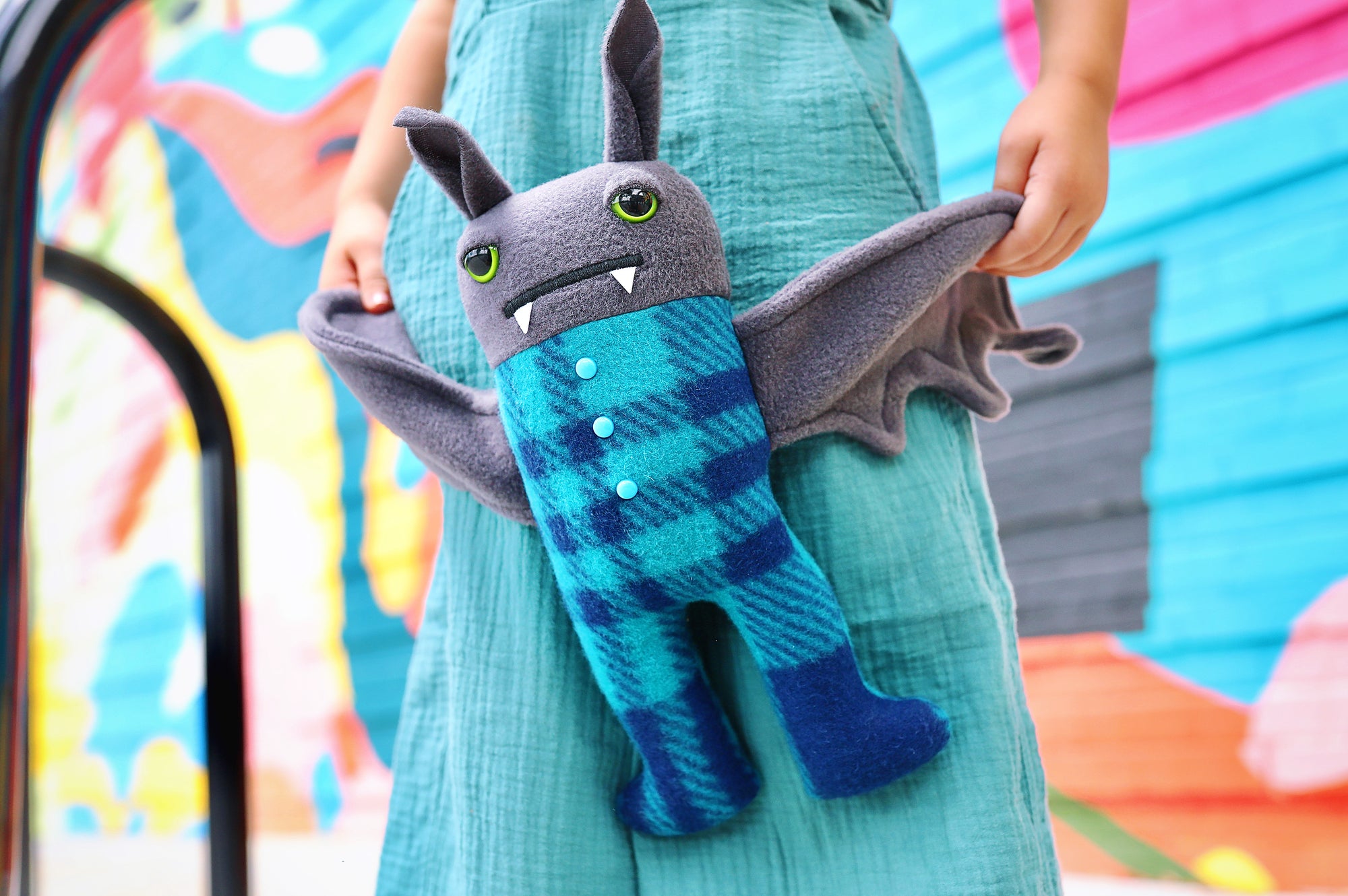 Girl stands holding bat plush by wings. They are both wearing teal and there is a colorful painted baclground.