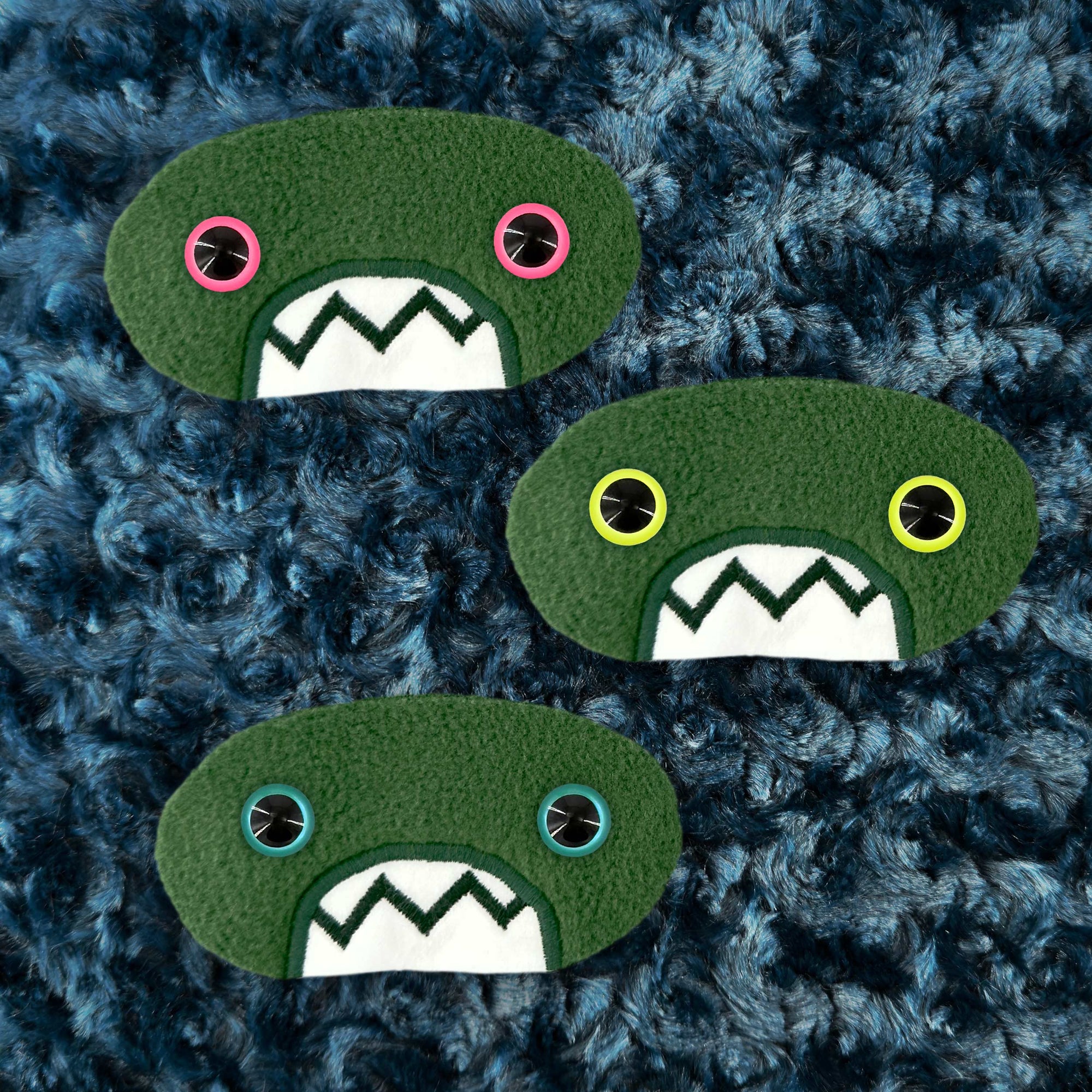 Three plush, green, oval-shaped patches with jagged mouths and colorful eyes are set against a textured, dark blue background resembling fur.