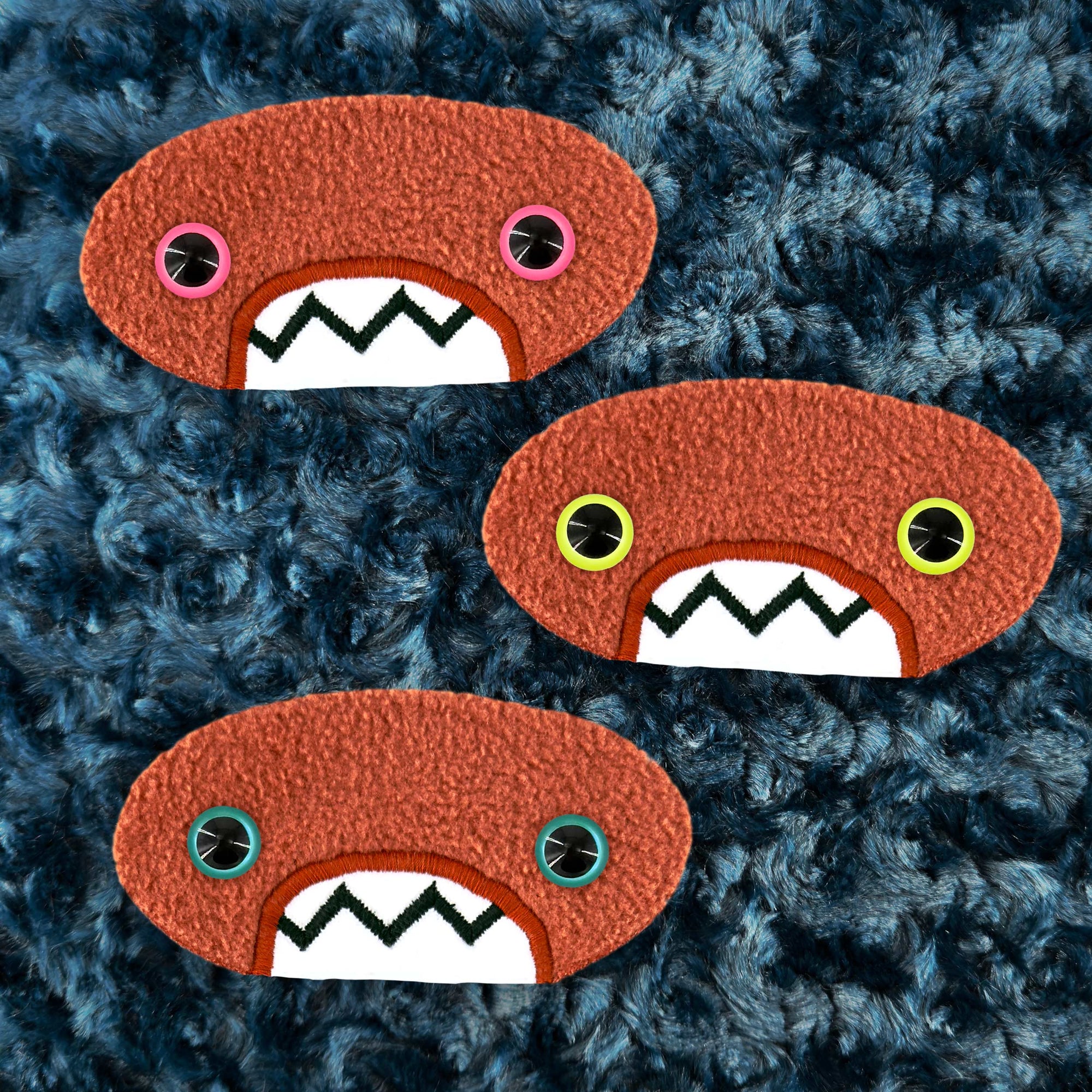 Three plush, orange, oval-shaped patches with jagged mouths and colorful eyes are set against a textured,  dark blue background resembling fur.