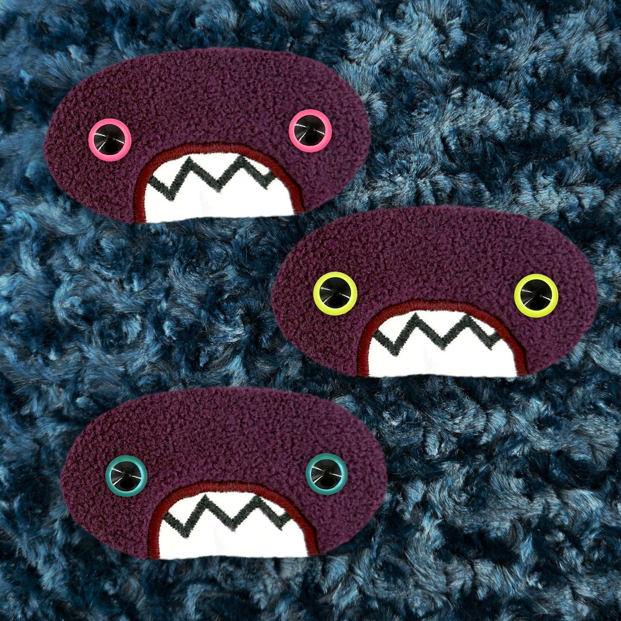 Three plush, purple, oval-shaped patches with jagged mouths and colorful eyes are set against a textured, dark blue background resembling fur.