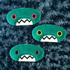 Three plush, teal, oval-shaped patches with jagged mouths and colorful eyes are set against a textured, dark blue background resembling fur.