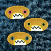 Three plush, mustard colored oval-shaped patches with jagged mouths and colorful eyes are set against a textured, dark blue background resembling fur.