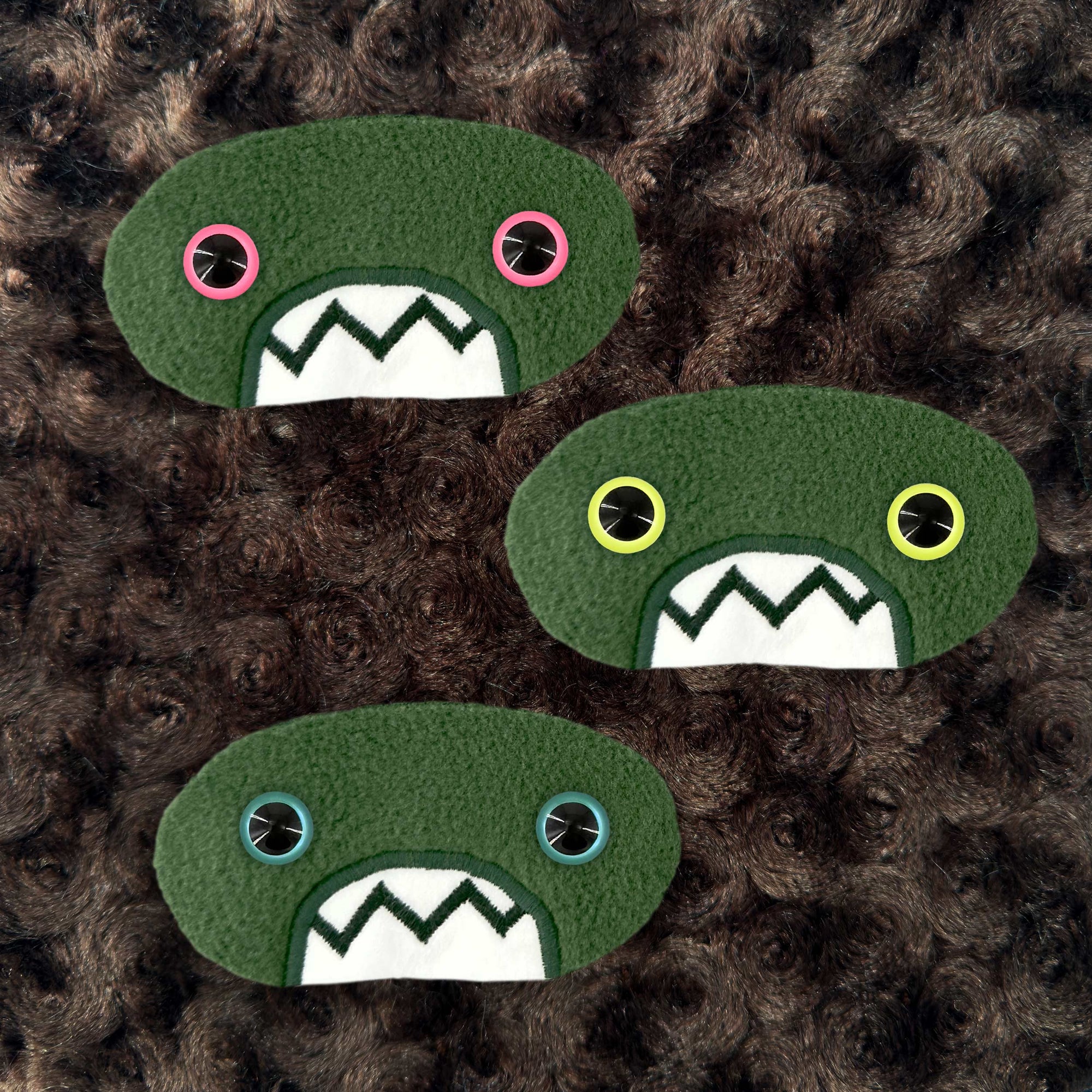 Three plush, green, oval-shaped patches with jagged mouths and colorful eyes are set against a textured, brown background resembling fur.