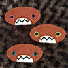 Three plush, orange, oval-shaped patches with jagged mouths and colorful eyes are set against a textured, brown background resembling fur.