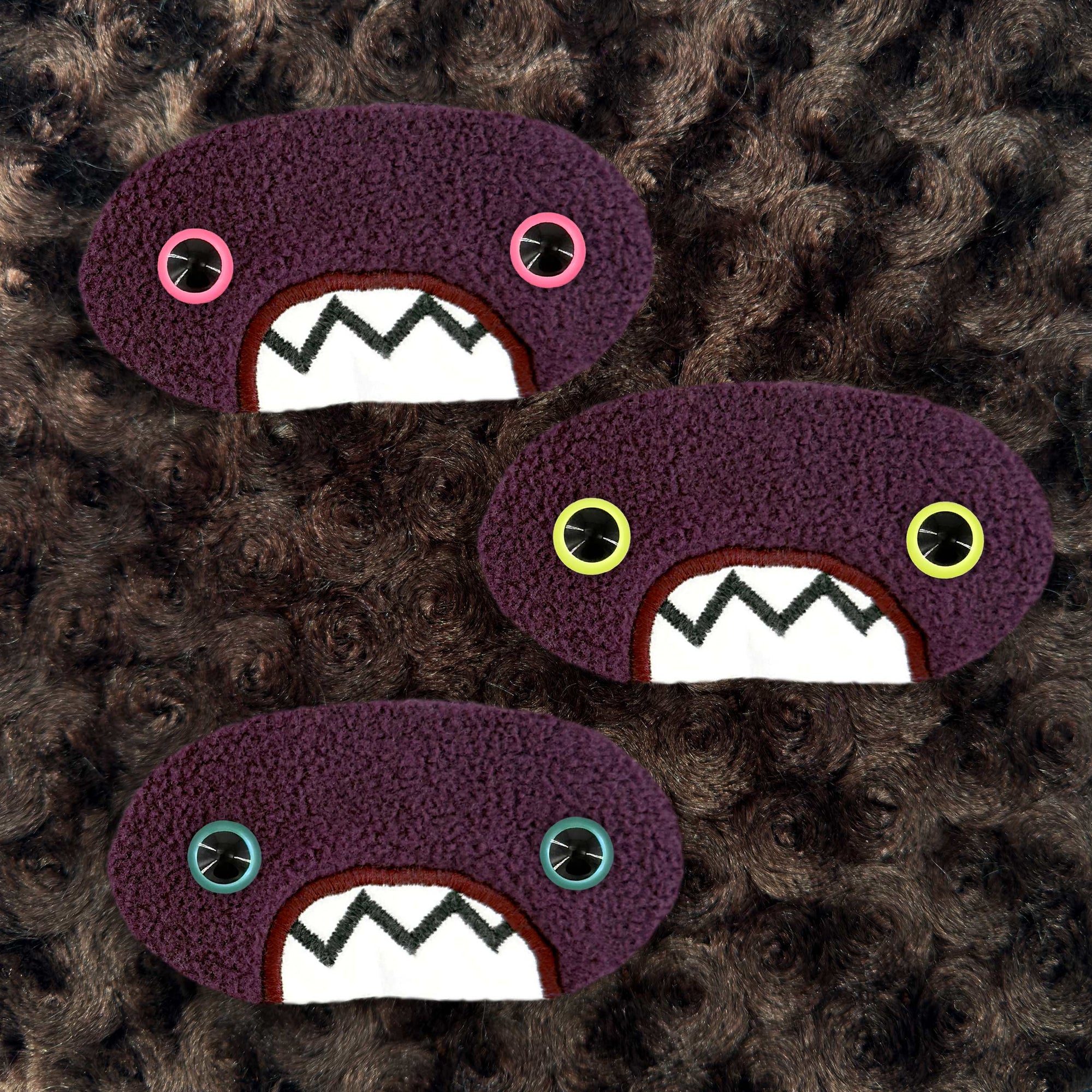 Three plush, purple, oval-shaped patches with jagged mouths and colorful eyes are set against a textured, brown background resembling fur.