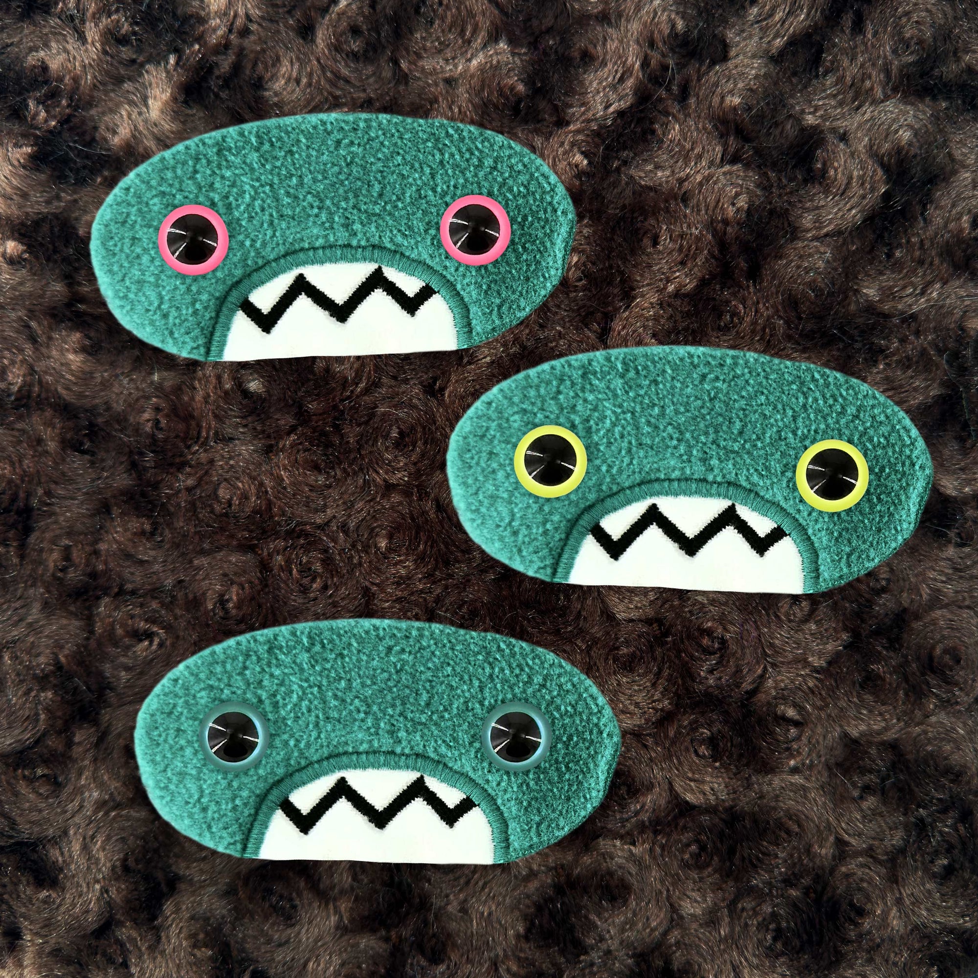 Three plush, teal, oval-shaped patches with jagged mouths and colorful eyes are set against a textured, brown background resembling fur.