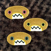 Three plush, mustard colored oval-shaped patches with jagged mouths and colorful eyes are set against a textured, brown background resembling fur.