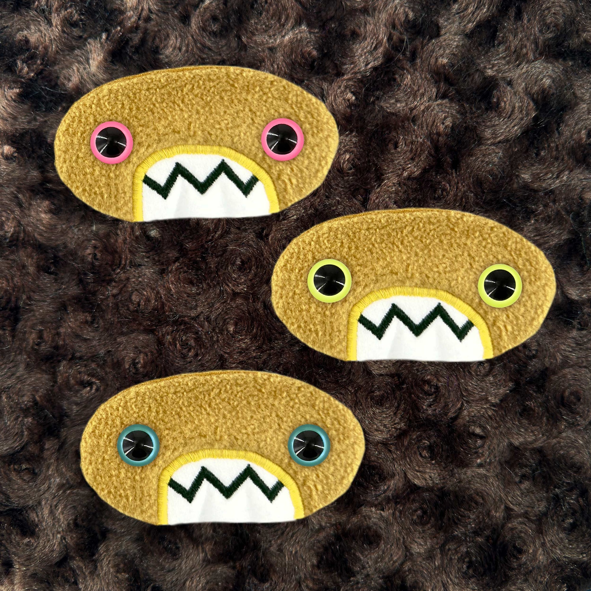 Three plush, mustard colored oval-shaped patches with jagged mouths and colorful eyes are set against a textured, brown background resembling fur.