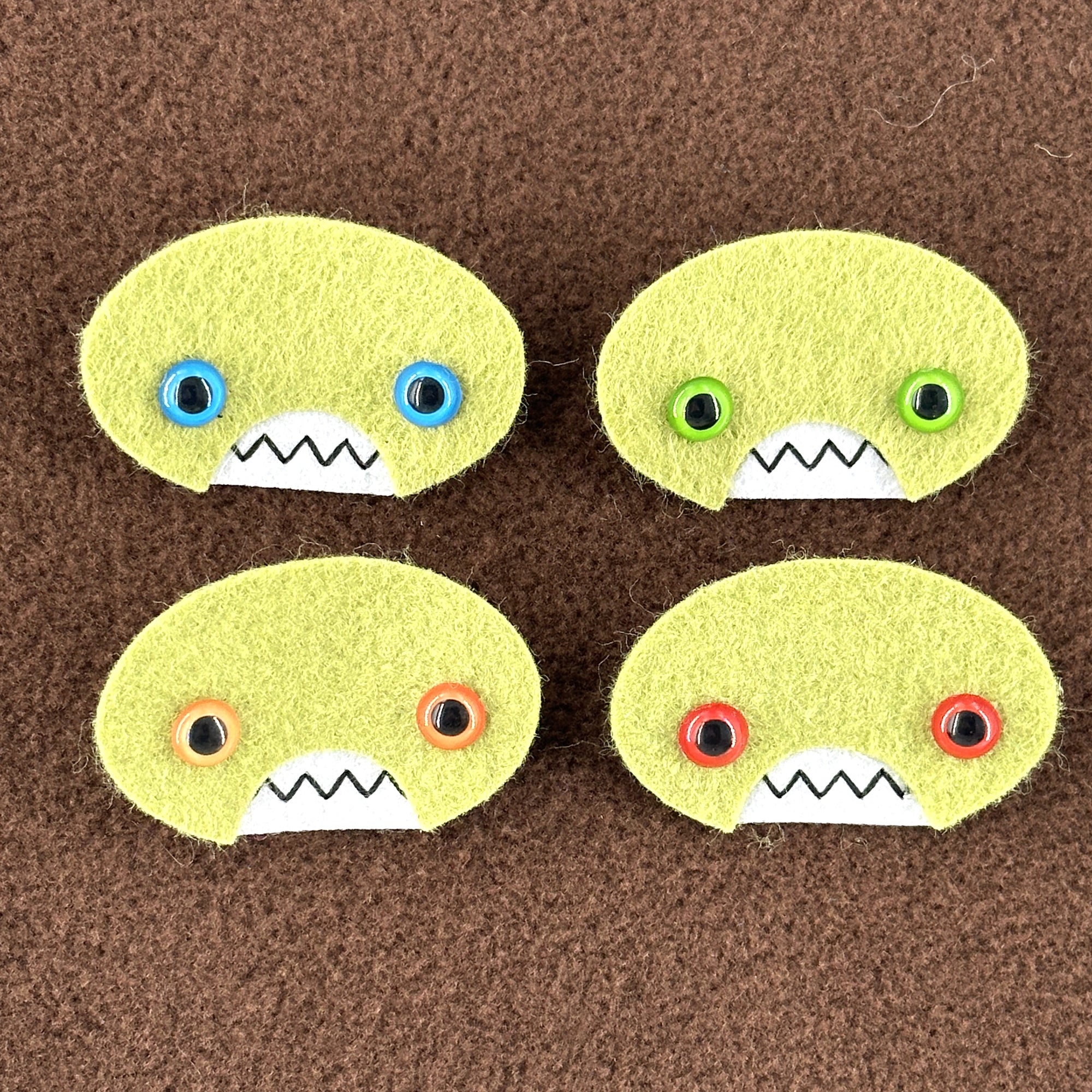 Four oval shaped green faces with different eye colors and pointed teeth sit atop a brown fleece background.