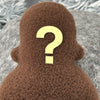 A small brown fleece figure with a question mark where its face should be stands in front of white foliage.