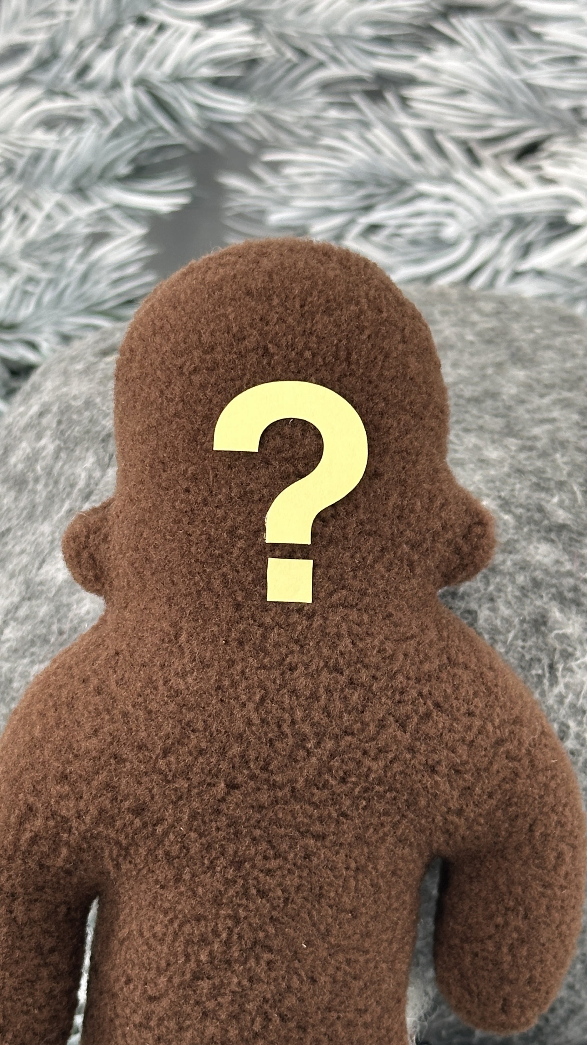 A small brown fleece figure with a question mark where its face should be stands in front of white foliage.