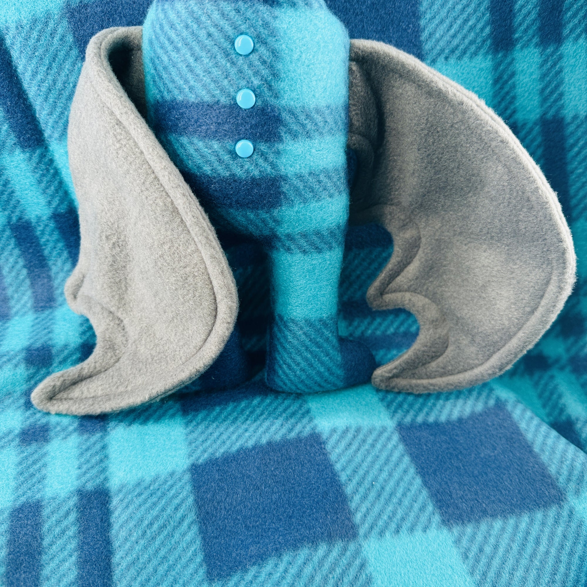 A plush bat wearing blue plaid pajamas sits a top a background of the same plaid fleece.