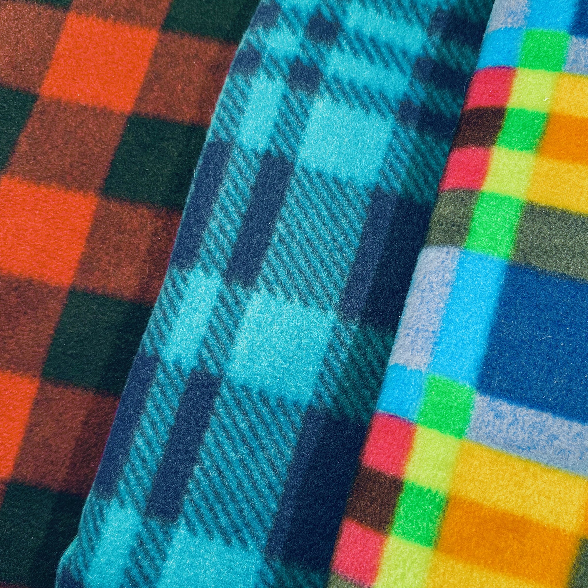 Close-up of three cozy plaid fabric patterns. The left side displays a red and brown plush fleece plaid, the middle presents a blue and teal design, while the right showcases a multicolored blend of blue, green, red, yellow, and gray.