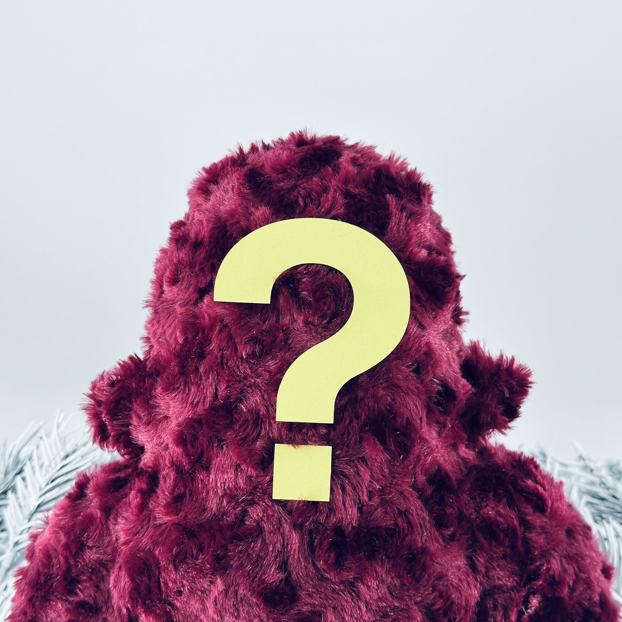 A fluffy  burgundy woolly yeti stands with their back to the camera, featuring a large yellow question mark. The pale, blurred background hints at white and gray accents resembling feathers or leaves, adding to the allure of this unique plush ensemble.