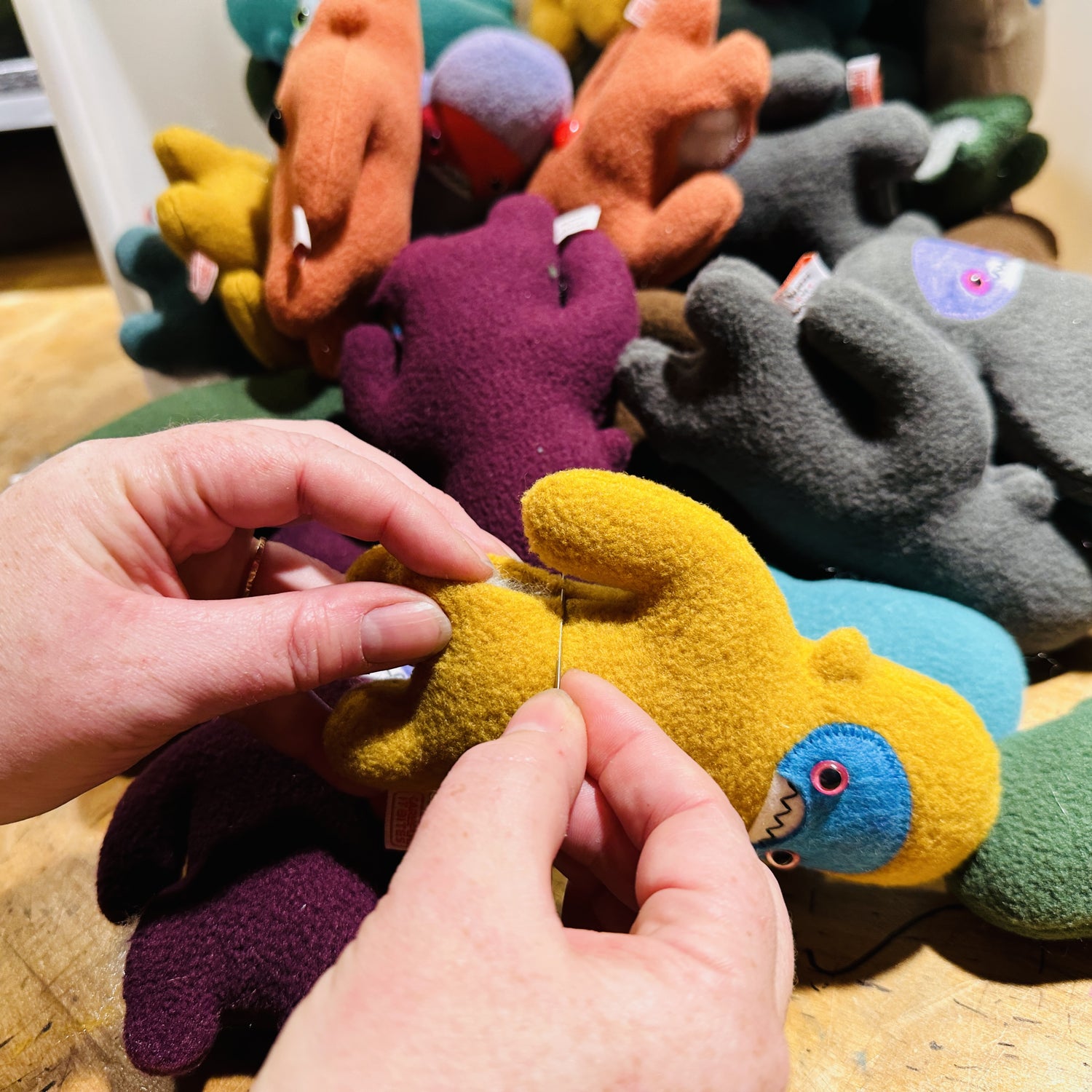 Hands sewing the side of a yellow fleece figure with a pikle of different colors creatures in the background.