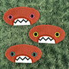 Three plush, orange, oval-shaped patches with jagged mouths and colorful eyes are set against a textured,  sage background resembling fur.