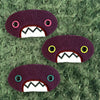 Three plush, purple, oval-shaped patches with jagged mouths and colorful eyes are set against a textured, sage background resembling fur.