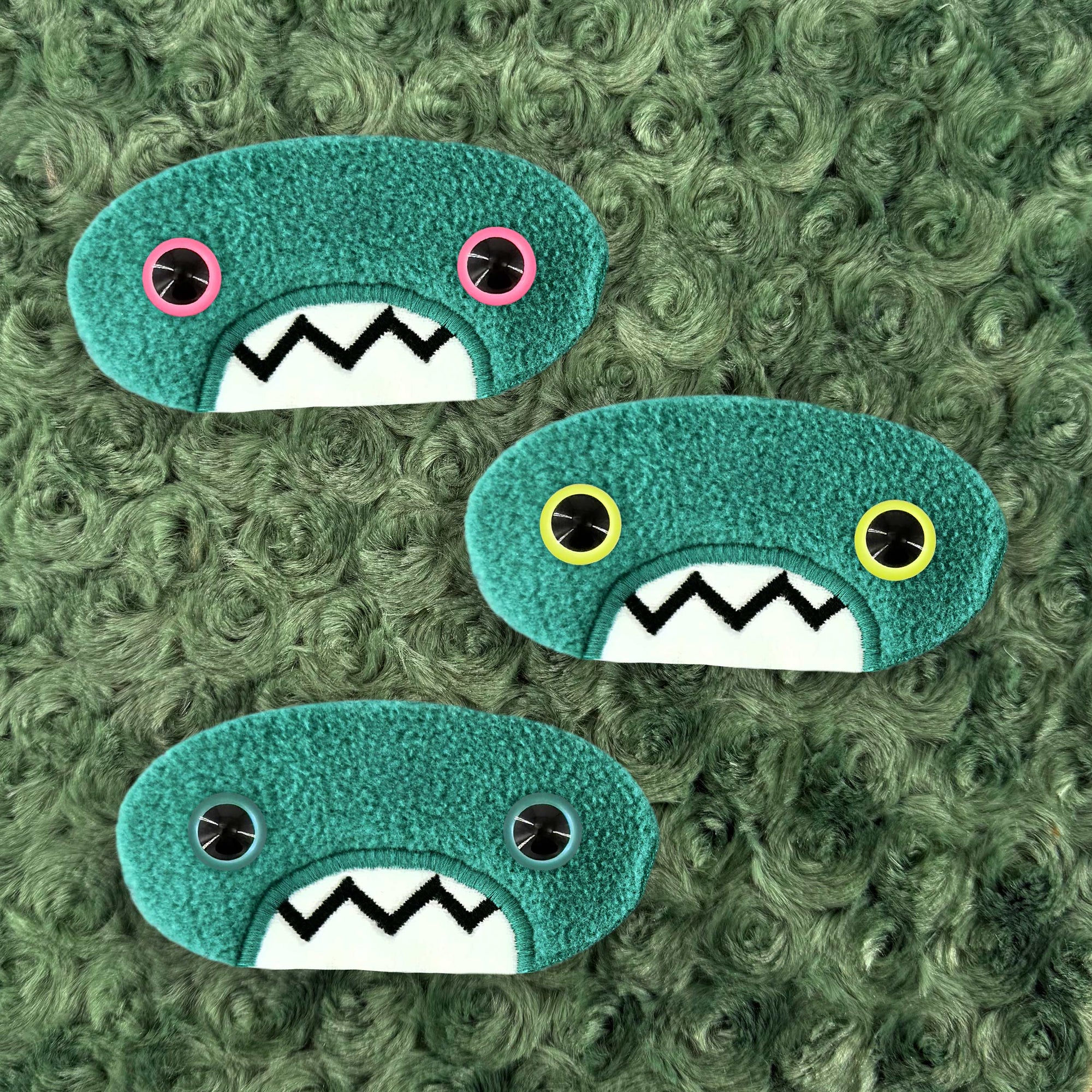 Three plush, teal, oval-shaped patches with jagged mouths and colorful eyes are set against a textured, sage background resembling fur.