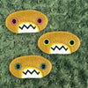 Three plush, mustard colored oval-shaped patches with jagged mouths and colorful eyes are set against a textured, sage green background resembling fur.