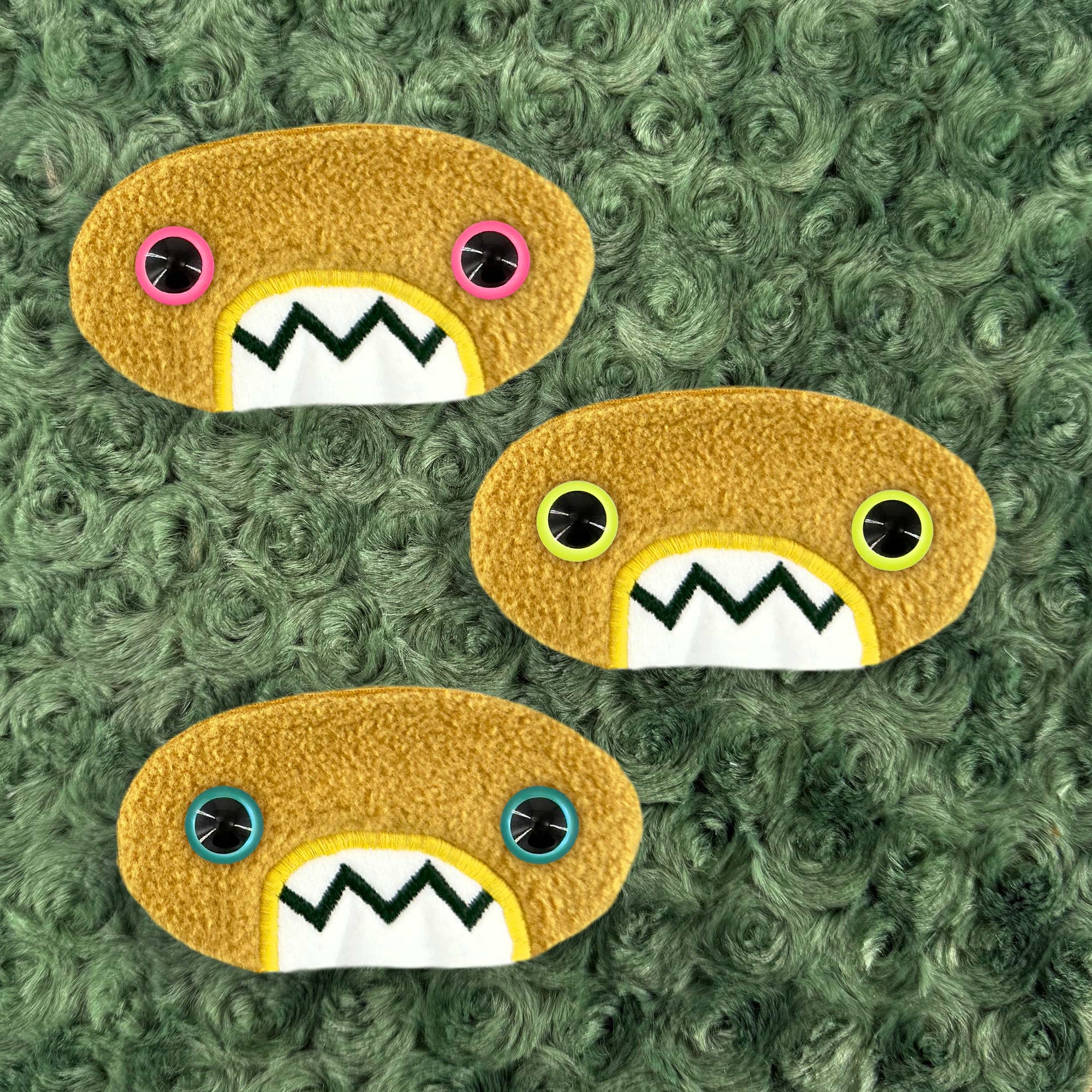 Three plush, mustard colored oval-shaped patches with jagged mouths and colorful eyes are set against a textured, sage green background resembling fur.