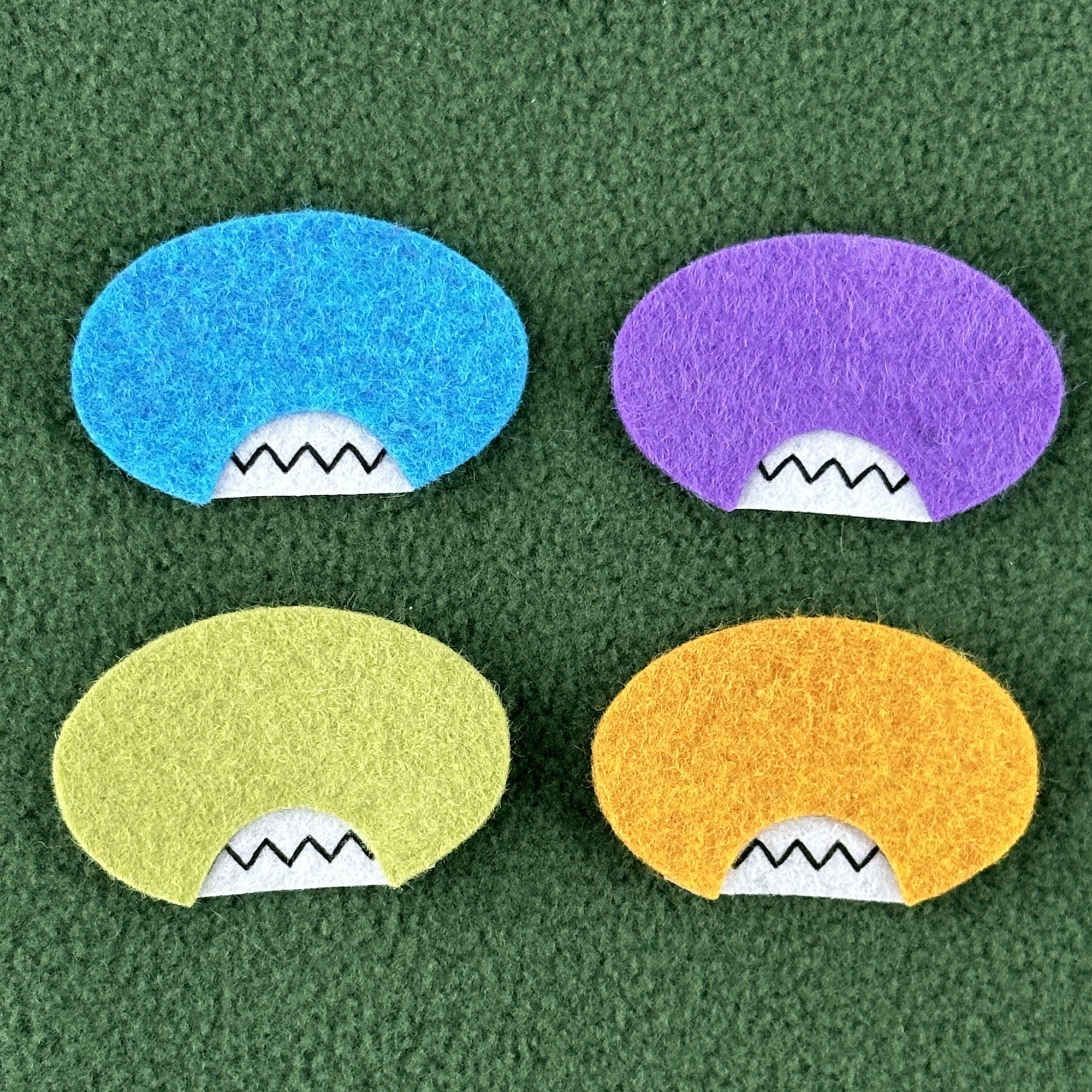 Four oval shaped faces in different colors and pointed teeth sit atop a green fleece background.