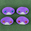 Four oval shaped purple faces with different eye colors and pointed teeth sit atop a green fleece background.