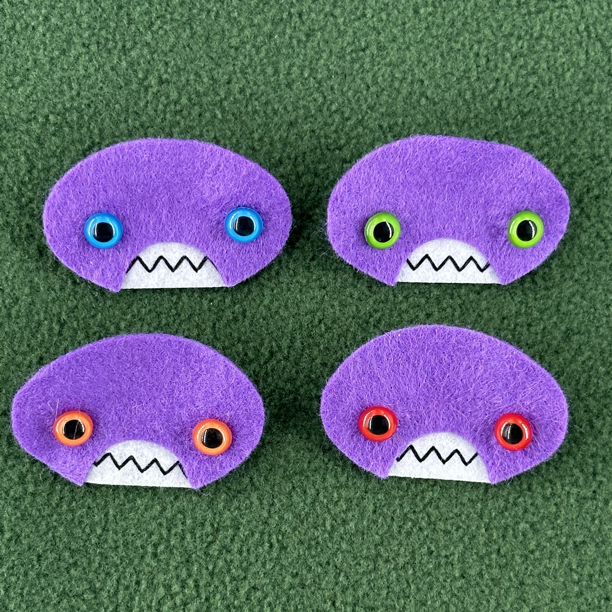 Four oval shaped purple faces with different eye colors and pointed teeth sit atop a green fleece background.