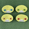 Four oval shaped green faces with different eye colors and pointed teeth sit atop a green fleece background.