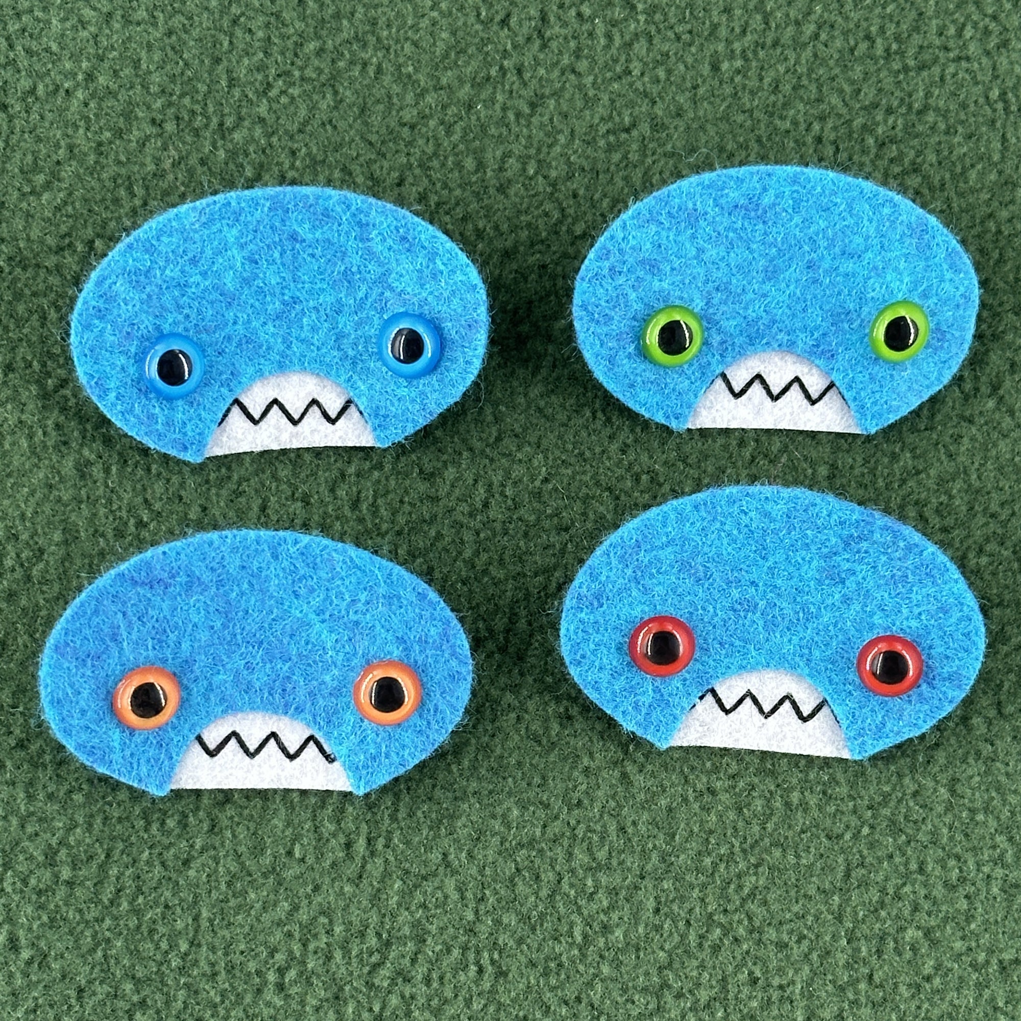 Four oval shaped teal faces with different eye colors and pointed teeth sit atop a green fleece background.