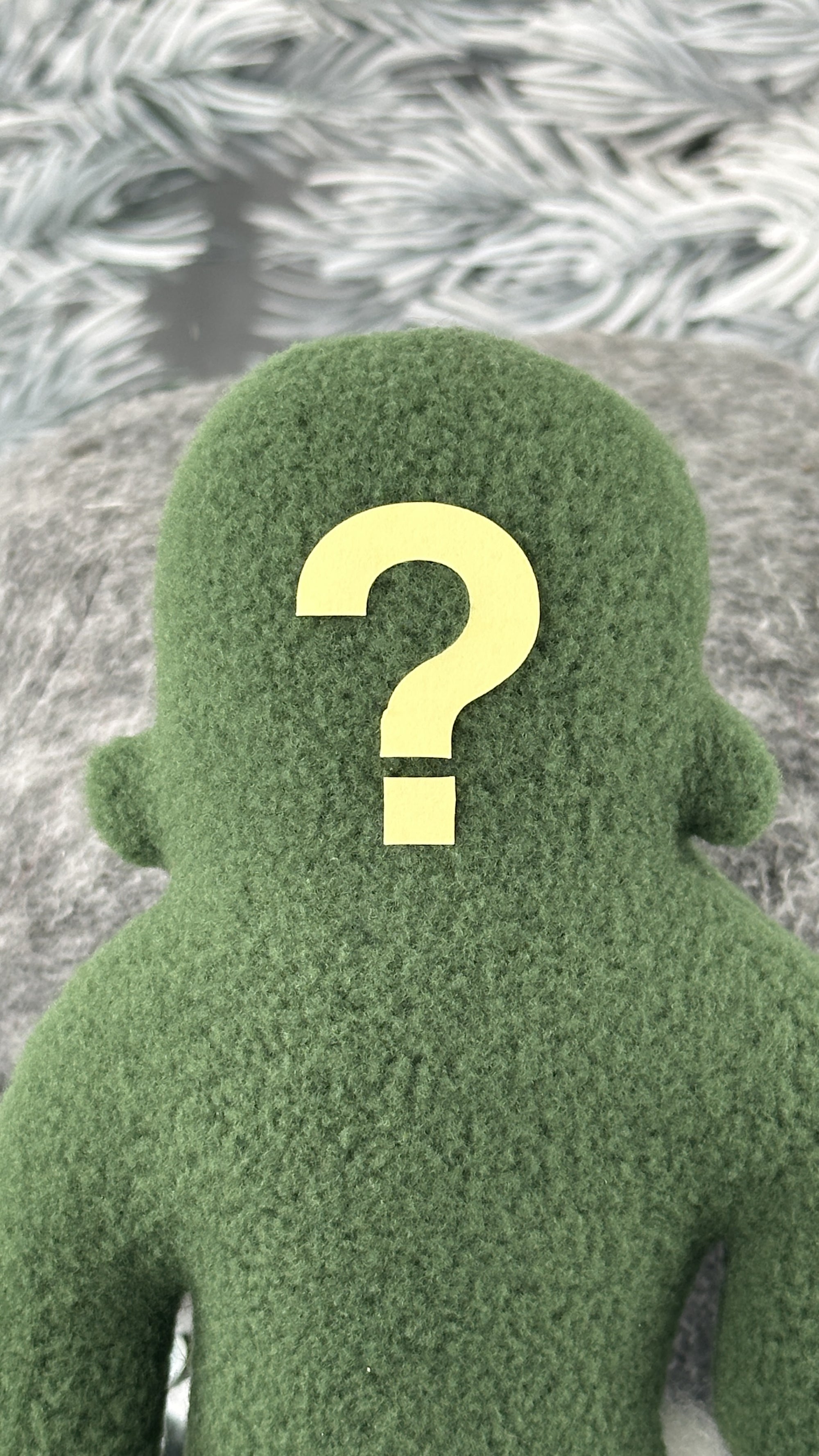 A small green fleece figure with a question mark where its face should be with blurred white background.