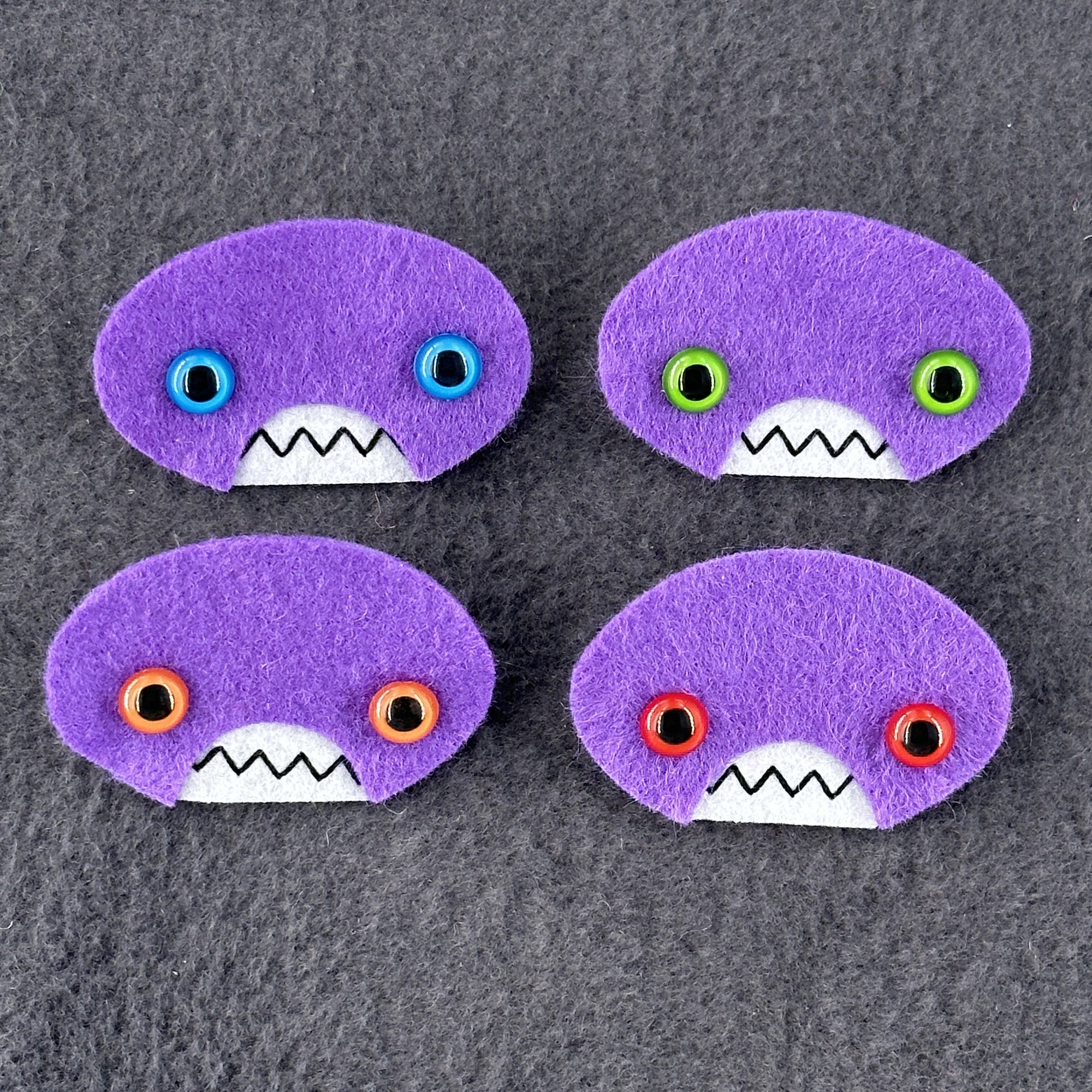 Four oval shaped purple faces with different eye colors and pointed teeth sit atop a grey fleece background.