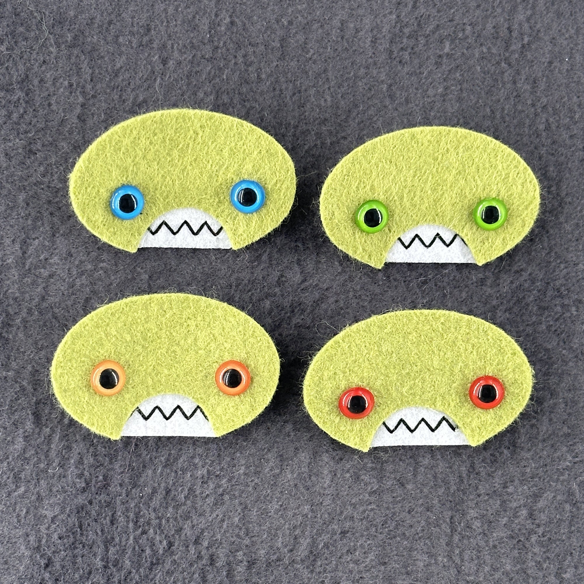 Four oval shaped green faces with different eye colors and pointed teeth sit atop a grey  fleece background.