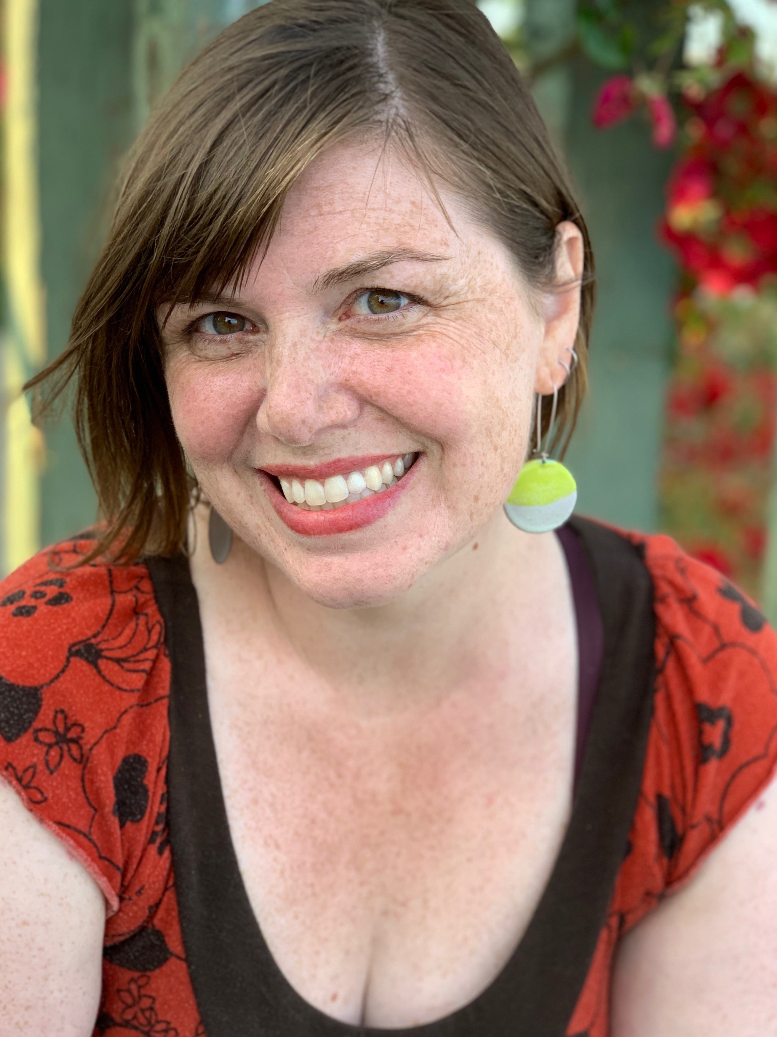 A smiling woman with short brown hair, wearing a red top with floral patterns and large round earrings in green and white, stands outdoors. Blurred greenery and flowers create a charming backdrop, echoing the warmth of cuddly companions as her dimples and freckles add to her cheerful demeanor.