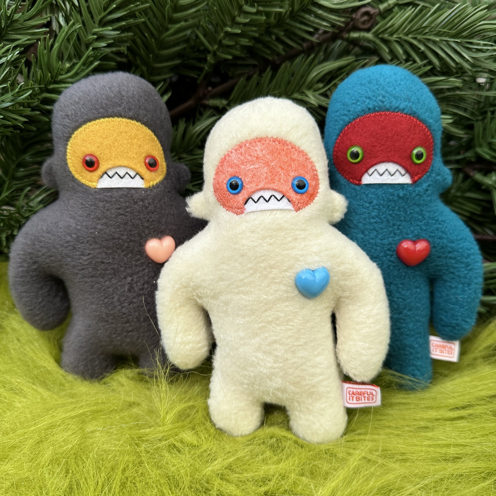 Group of three colorful plush sasquatch tand on fake grass with foliage behind.