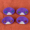 Four oval shaped purple faces with different eye colors and pointed teeth sit atop a orange fleece background.