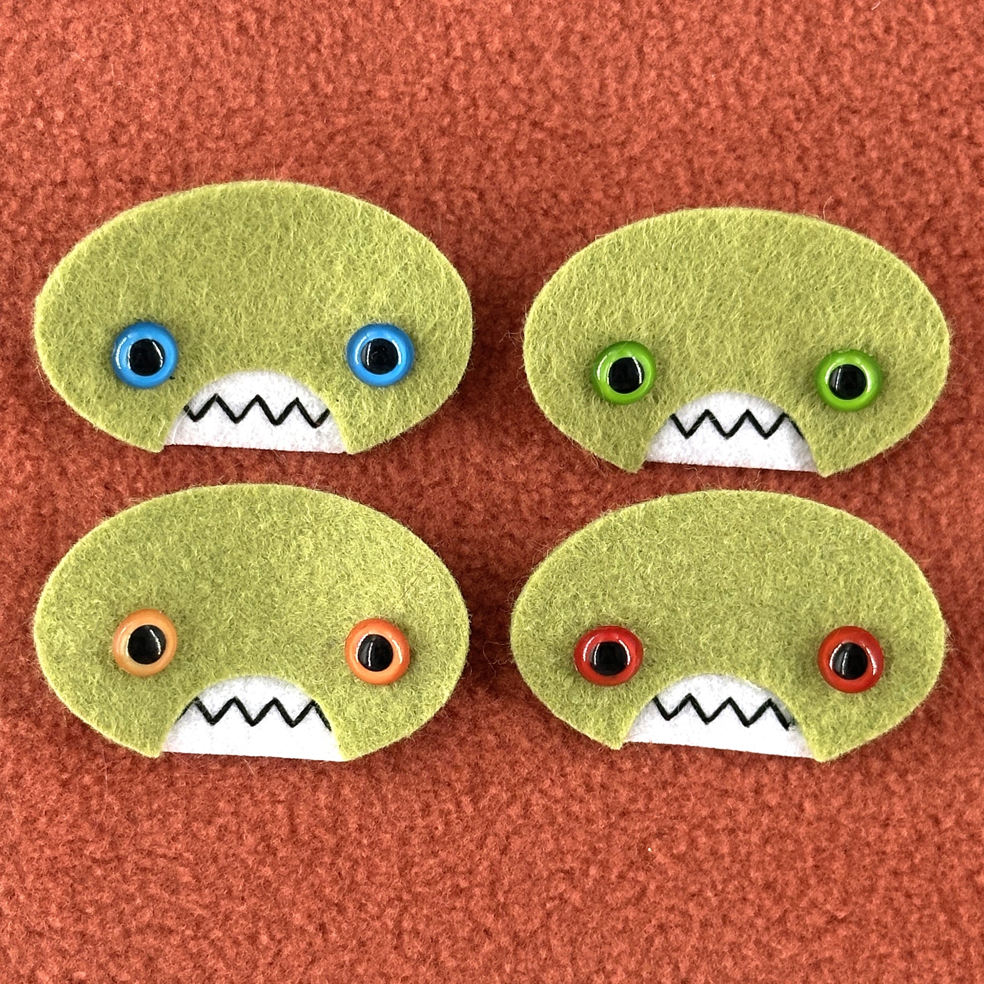 Four oval shaped green faces with different eye colors and pointed teeth sit atop an orange fleece background.