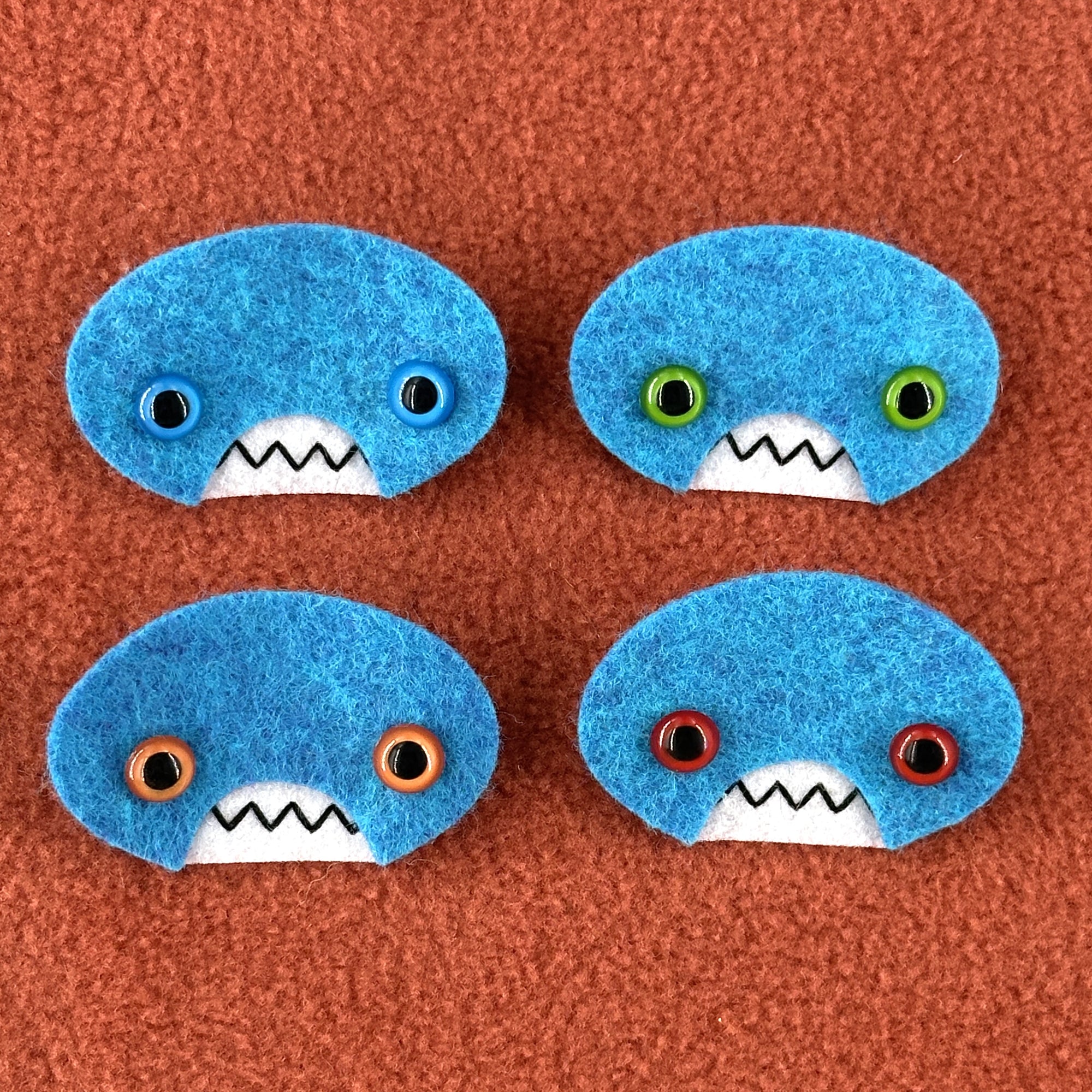 Four oval shaped teal faces with different eye colors and pointed teeth sit atop an orange fleece background.